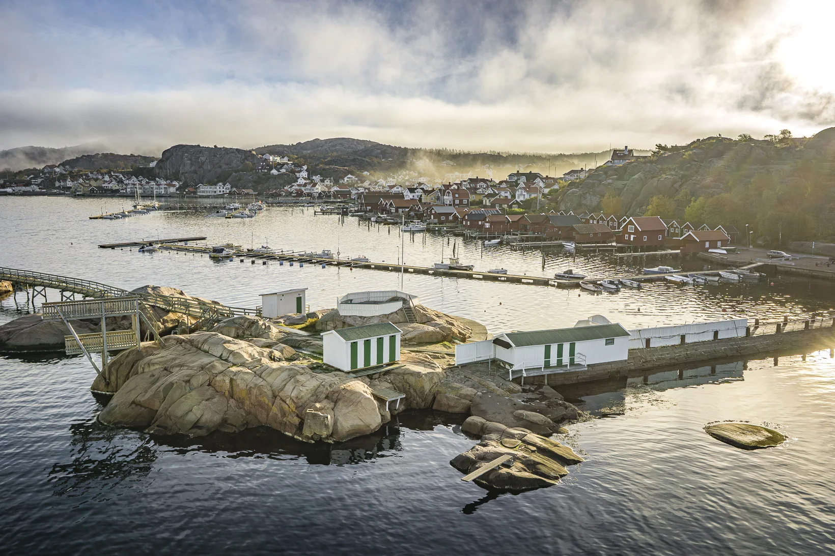Badholmarna im Gegenlicht, mit den Badehütten, dem Steg und den Bootsstegen, dahinter der Ort Bovallstrand